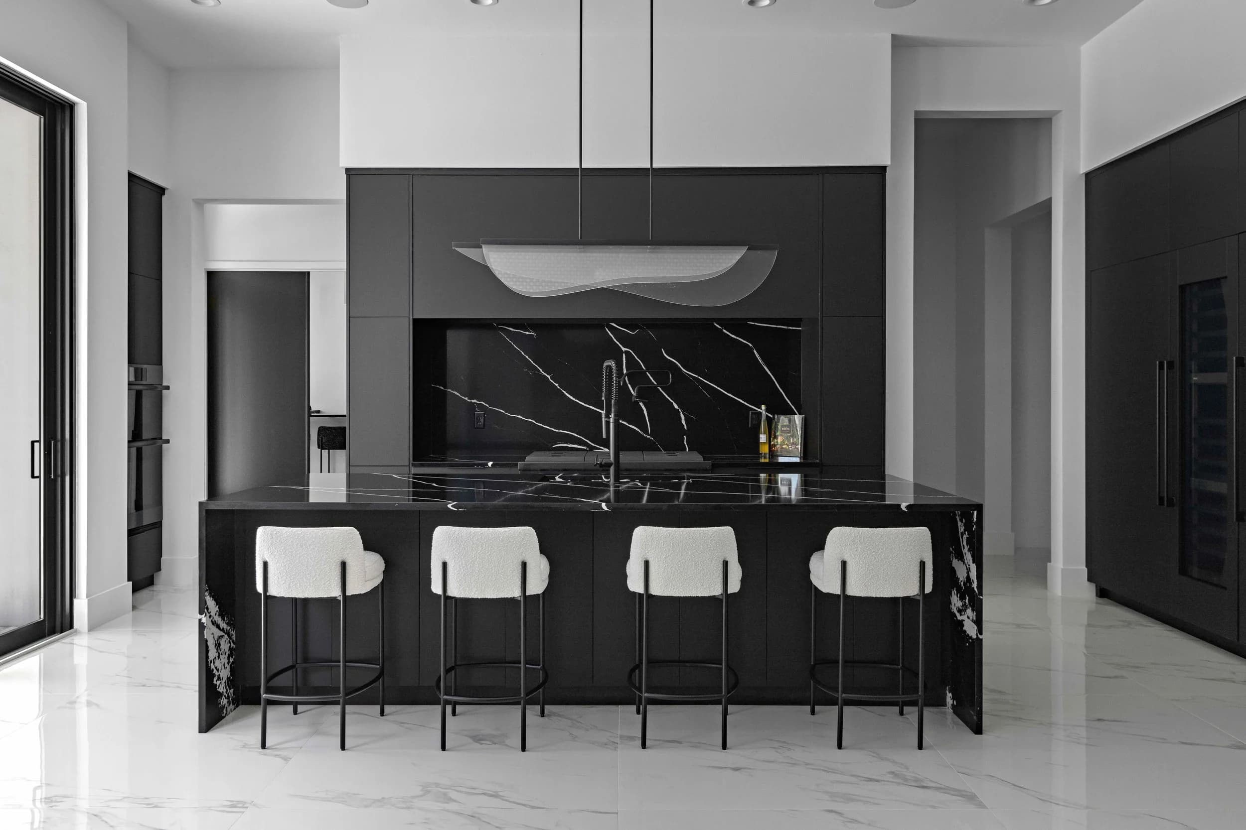 Monochrome kitchen with a black marble island, white stools, and white porcelain floor